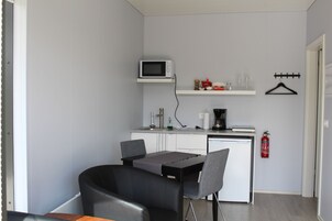 Cocina compacta con microondas, cafetera y un estante pequeño con diversos objetos. Hay una mesa de comedor con sillas y un extintor de incendios montado en la pared.