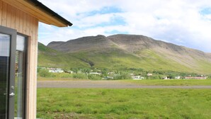 A wooden building with a glass door, a grassy field, and a mountain in the background. - Systrakot (Talknafjordur)