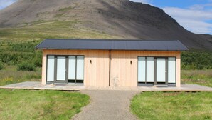 A modern building with a wooden exterior and large glass doors, situated in a grassy area with a mountain in the background. - Systrakot (Talknafjordur)