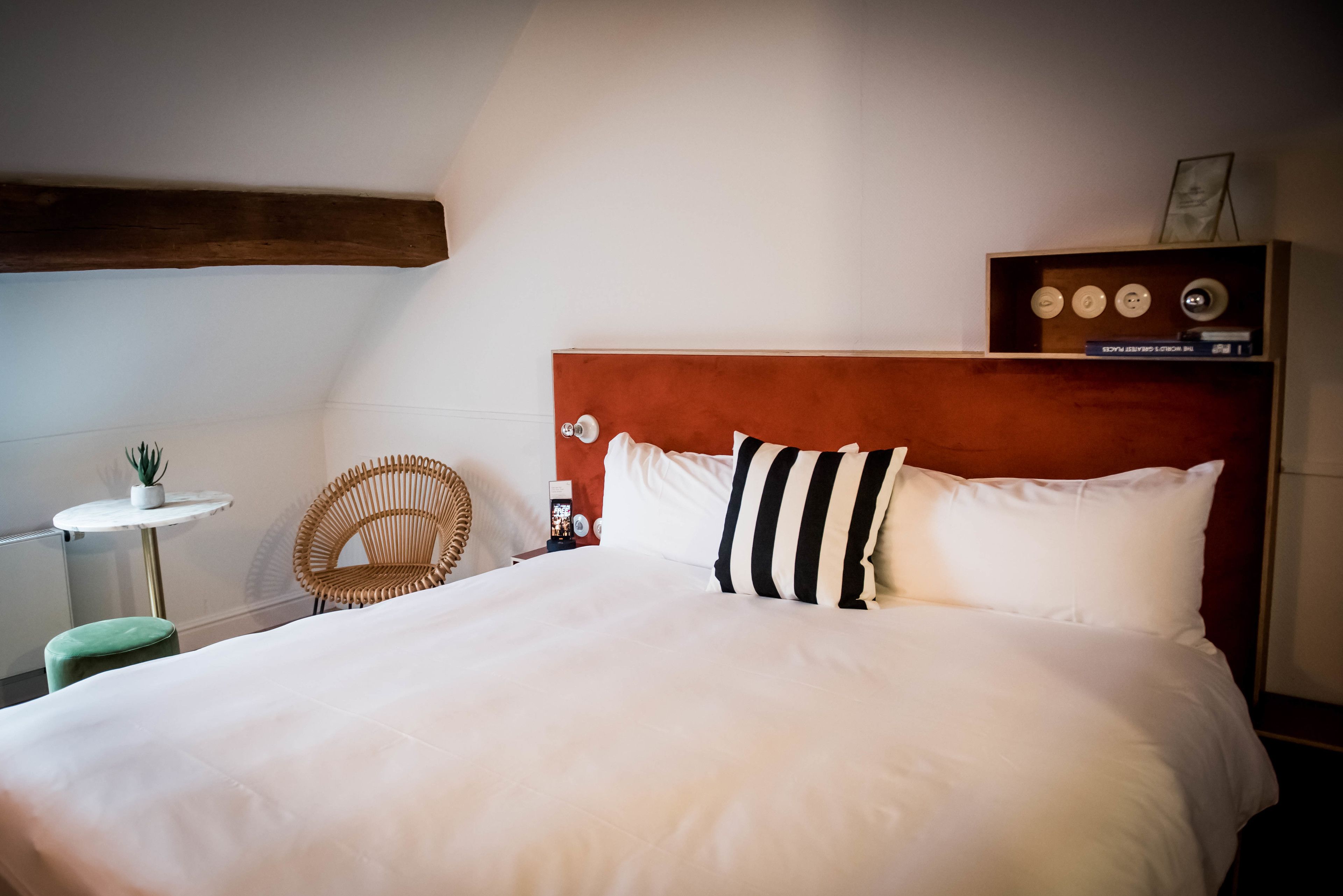A neatly made bed with white linens and a striped pillow, a wooden headboard, a small round table with a plant, and a wicker chair.