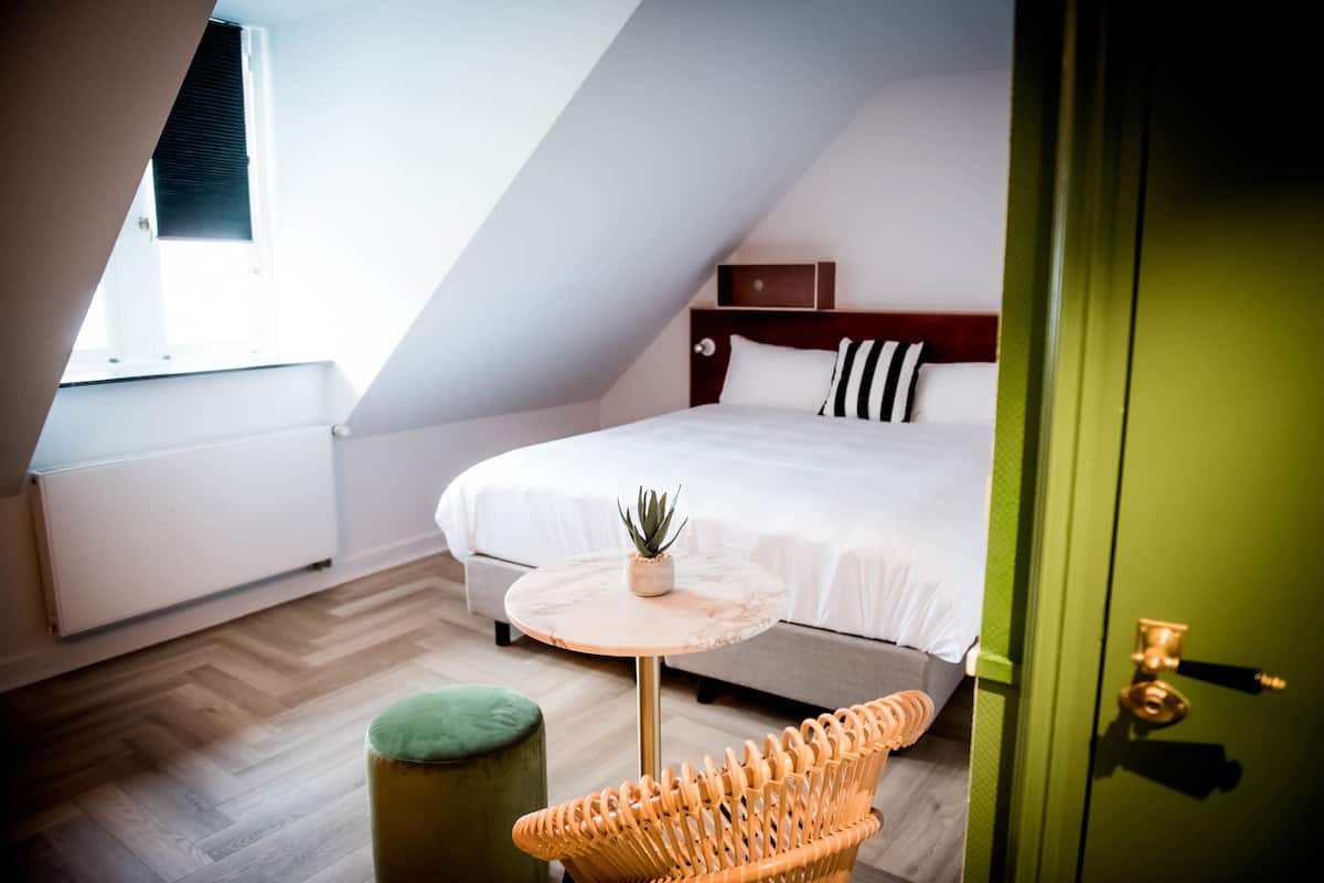 A bedroom with a bed, a small round table, a green ottoman, and a window with blinds.