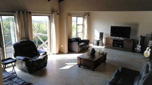 A spacious living room with a sofa, armchair, and coffee table. There is a television, a guitar, and a window with curtains. - Coastal Haven 4 (Gqeberha (Port Elizabeth))