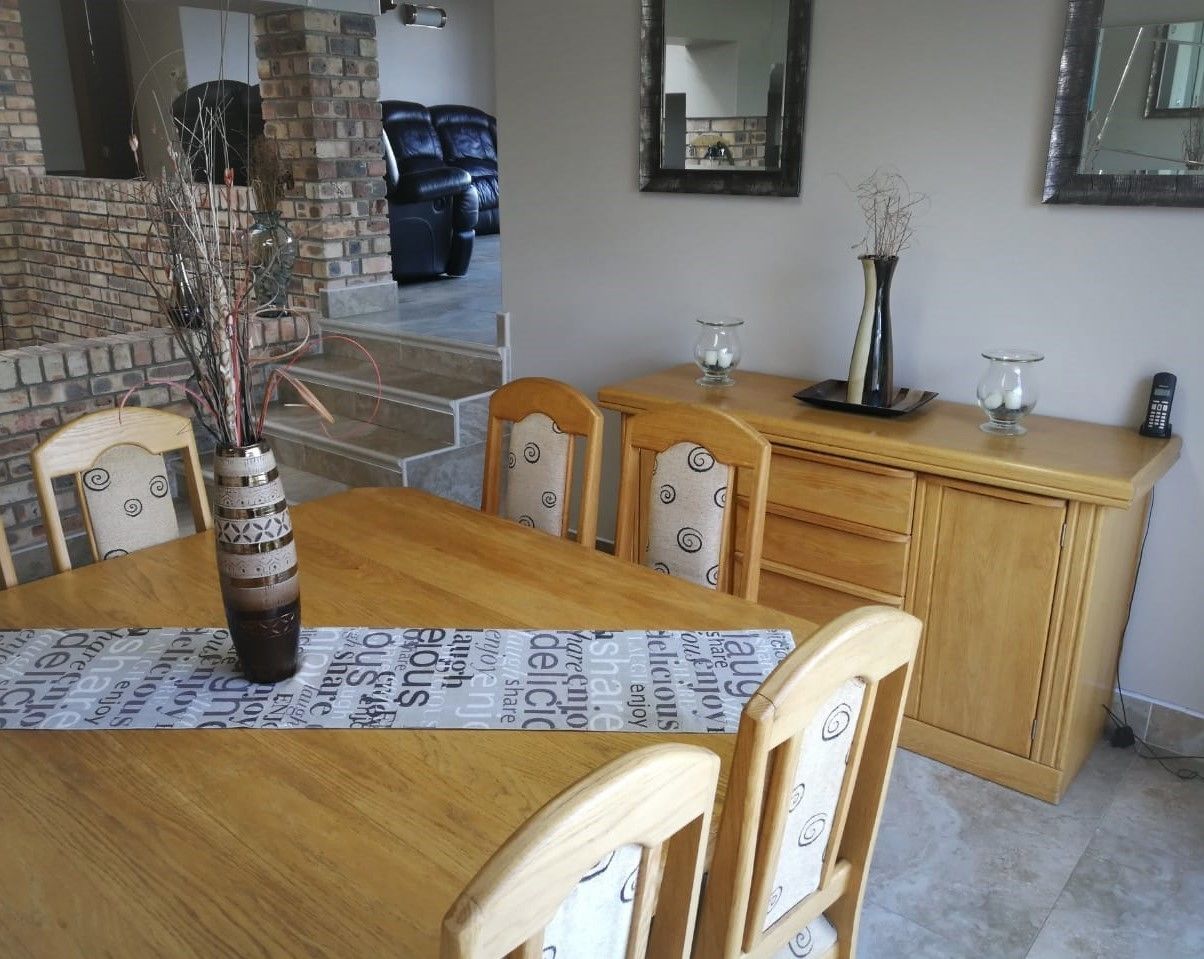 A dining area with a wooden table, chairs, and a vase with branches. There is a brick wall and a mirror on the wall.