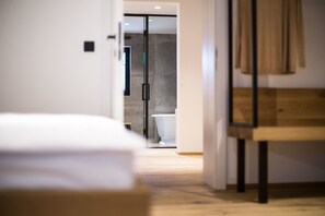 A modern hotel room with a bed, a wooden bench, and a view of the bathroom.