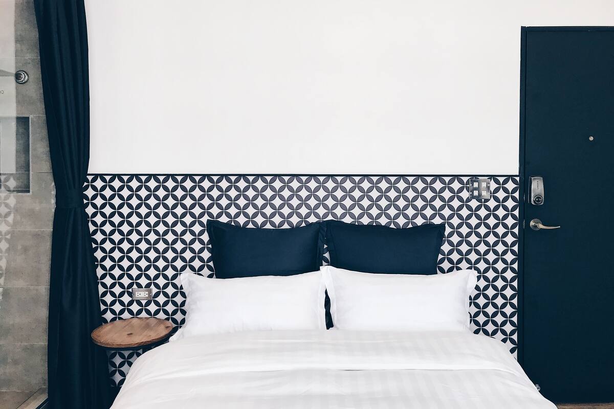 A bed with white bedding, two dark pillows, and a wooden nightstand with a white plate.