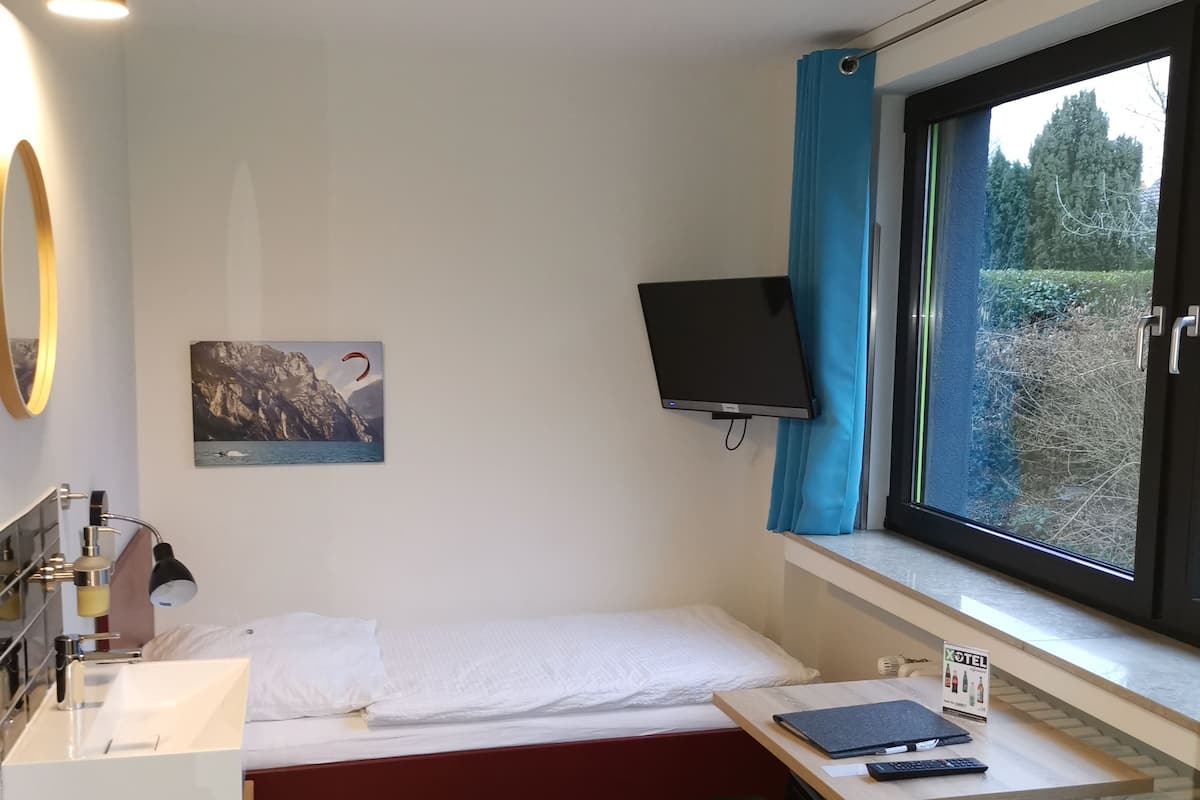 A compact hotel room with a bed, a desk, a computer, a TV, and a window with a view of trees.