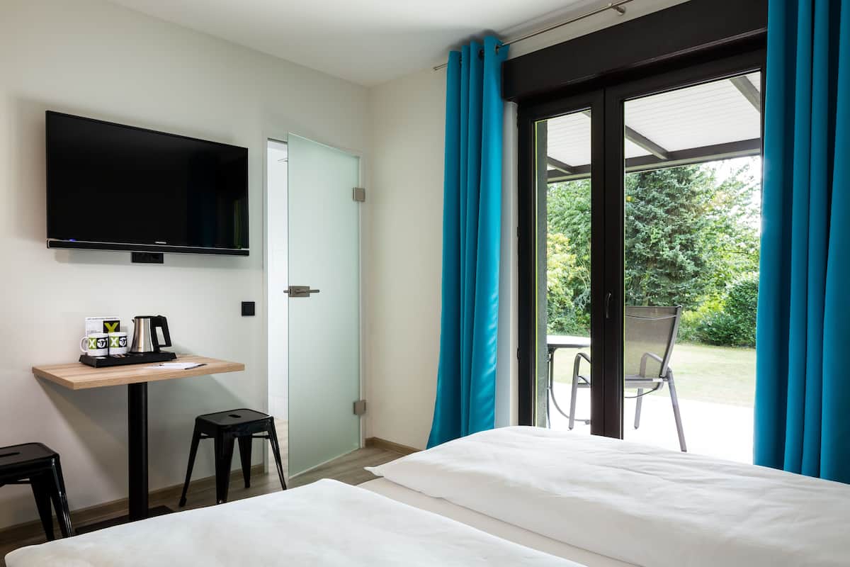 A hotel room with a bed, a TV mounted on the wall, a small table with a coffee maker, and a chair outside the window.