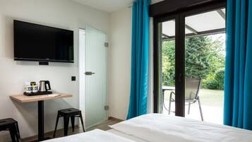 A hotel room with a bed, a TV mounted on the wall, a small table with a coffee maker, and a chair outside the window.