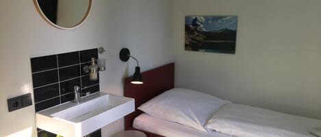 Una habitación pequeña y moderna con una cama individual, una mesita de noche blanca, un taburete pequeño, un espejo redondo, un lavamanos instalado en la pared y un cuadro de un paisaje.