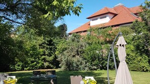 An outdoor seating area with tables and chairs, surrounded by lush greenery and a red-roofed building in the background. - Xotel (Xanten)