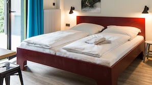 A bedroom with a wooden bed, white linens, a view through a glass door, and a blue curtain. - Xotel (Xanten)