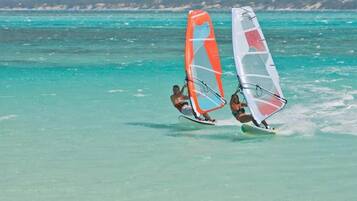 Dos personas practicando windsurf en aguas turquesas, con una costa montañosa al fondo.