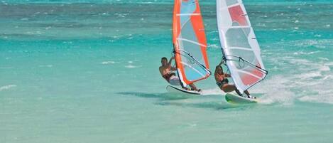 Two people windsurfing on turquoise water with a mountainous coastline in the background.
