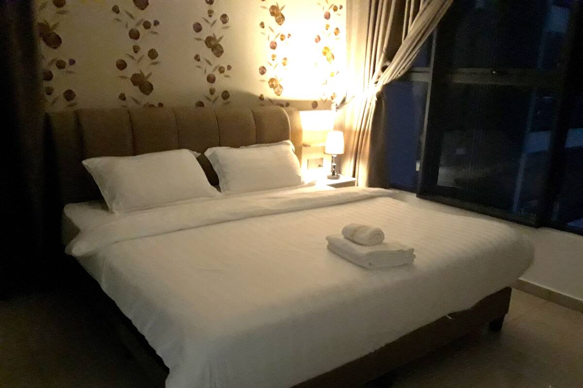 A neatly made bed with white linens and a rolled towel on top, a nightstand with a lamp, and a window with curtains.