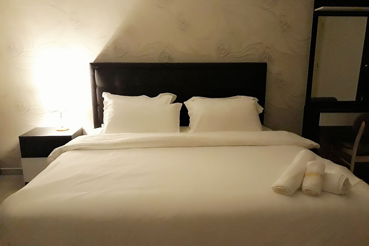 A neatly made bed with white linens and pillows, a dark headboard, and a nightstand with a lamp.
