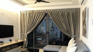 Ein modernes Wohnzimmer mit einer schwarzen Couch, einem Couchtisch und einem Fernseher an einer Marmorwand. Große Fenster mit Vorhängen bieten Blick auf die Stadtlandschaft.