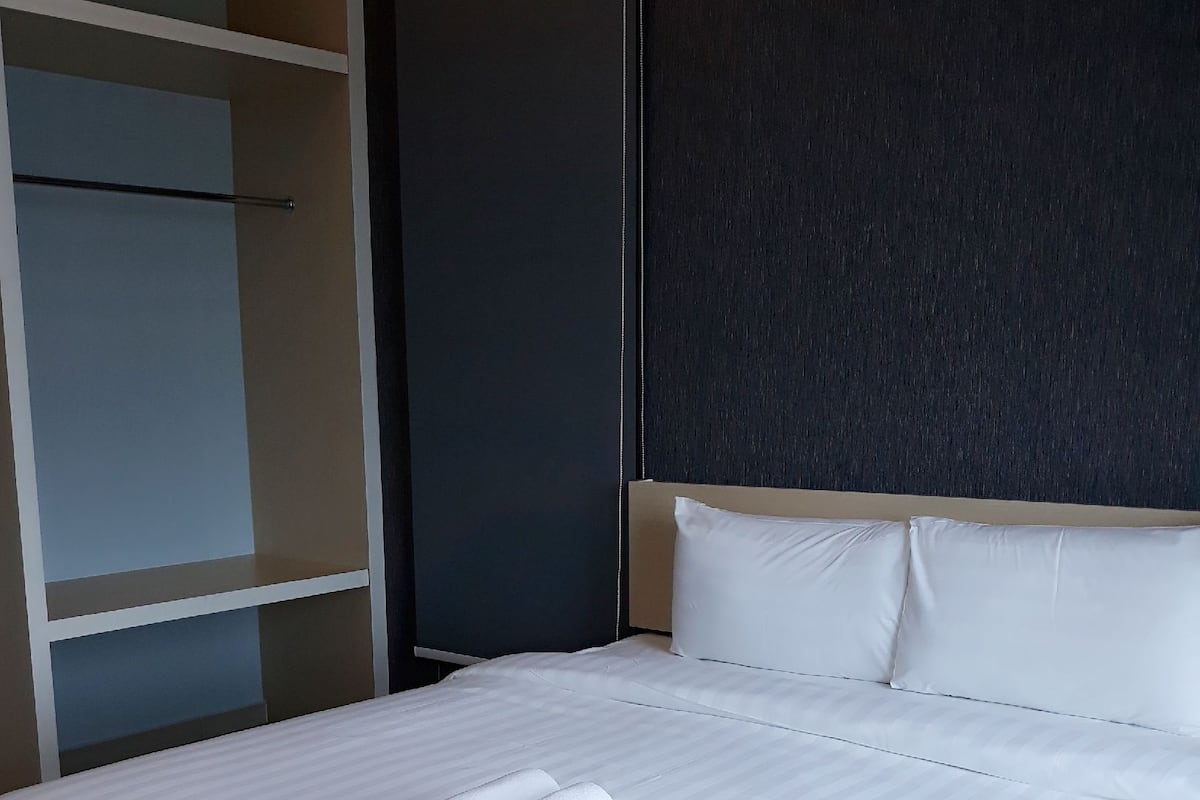 A neatly made bed with white linens and a rolled towel, a dark wall, and a partially visible wardrobe.