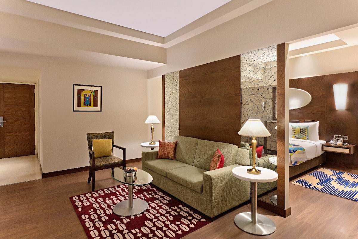 Une chambre d’hôtel avec un canapé vert, une chaise, une petite table, une lampe, un lit et un miroir.