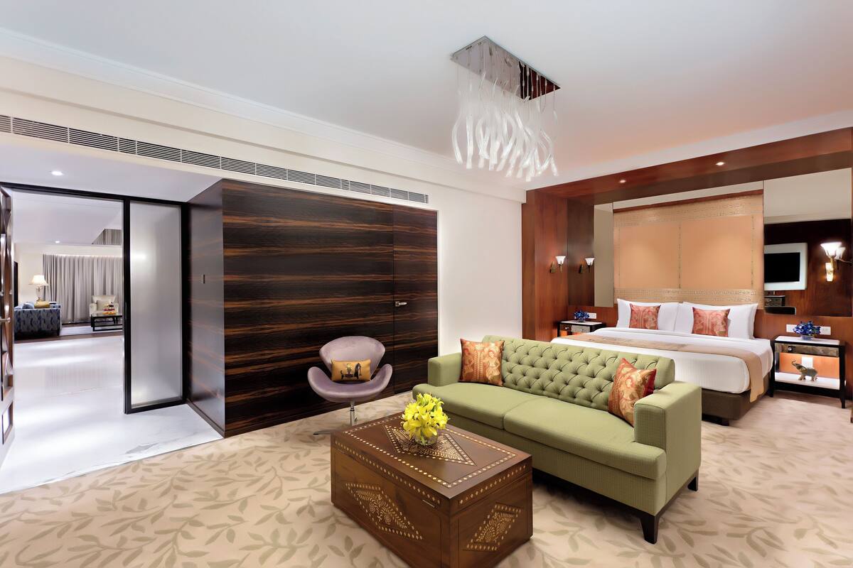 Une chambre d’hôtel moderne avec un canapé vert, une table basse en bois, un grand lit et un espace nuit séparé comprenant un bureau et une chaise.