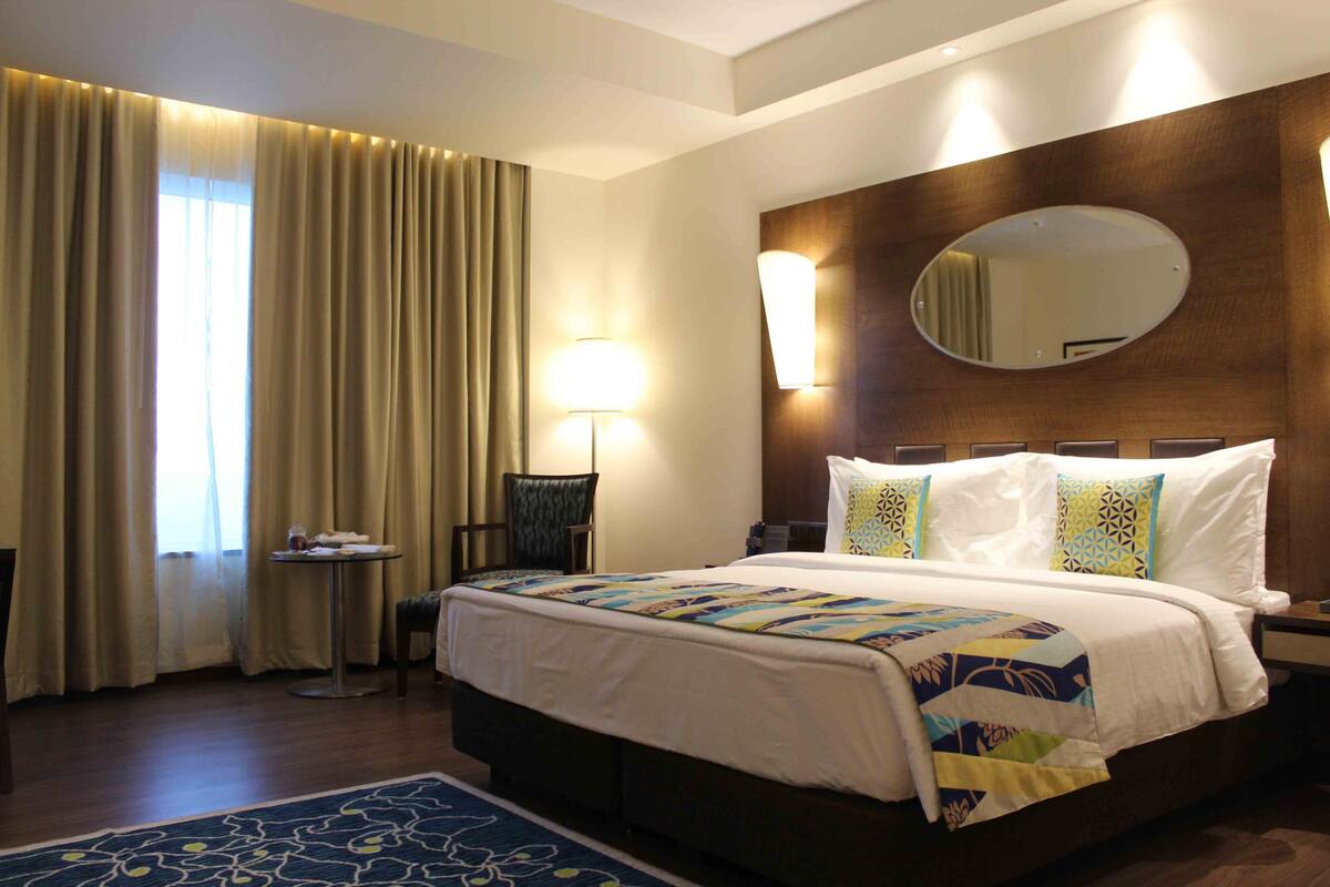 Une chambre d’hôtel avec un grand lit, un miroir rond, une table de chevet, une chaise et une fenêtre avec des rideaux.