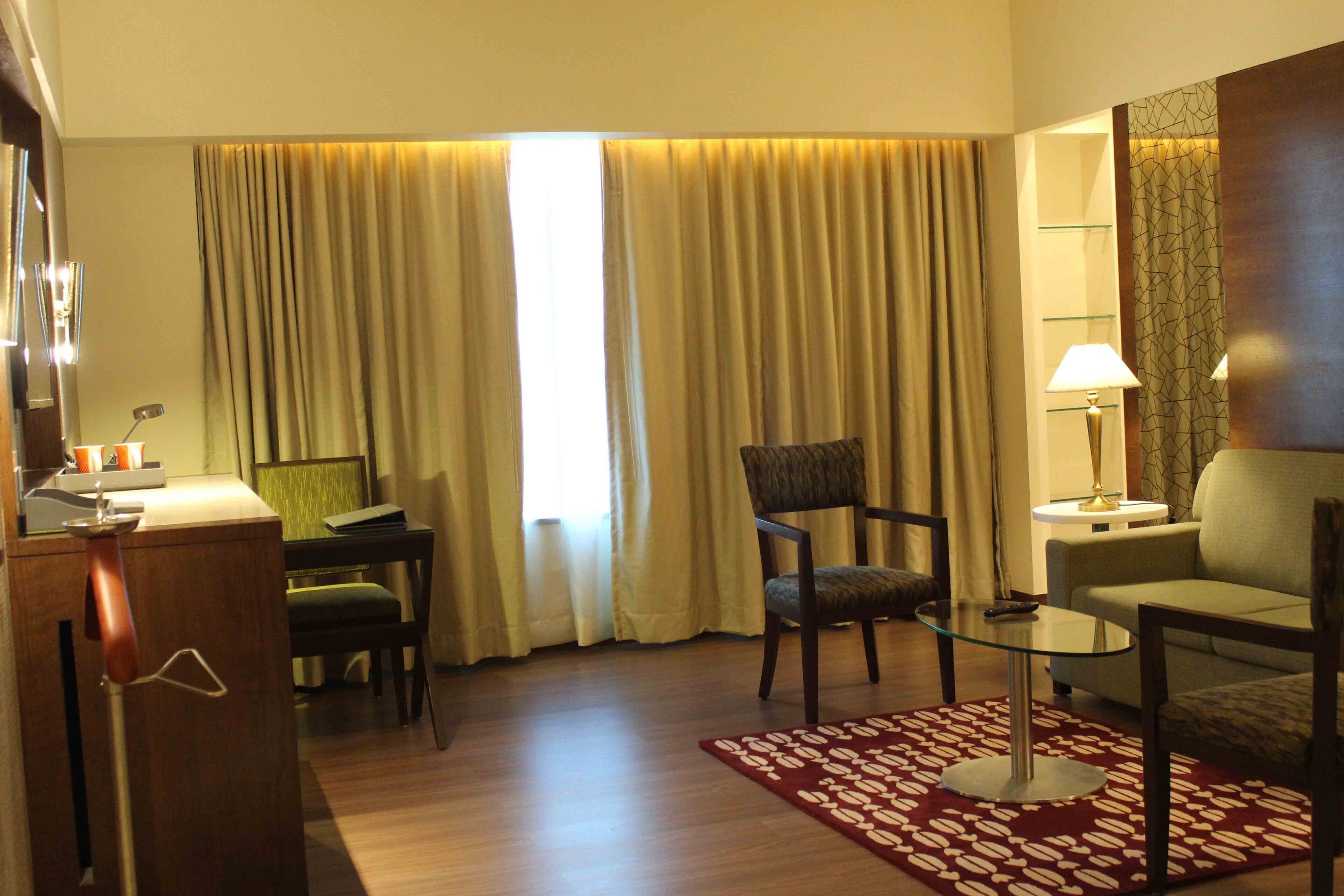 A hotel room with a wooden desk, a glass table, a chair, a sofa, and a patterned rug.
