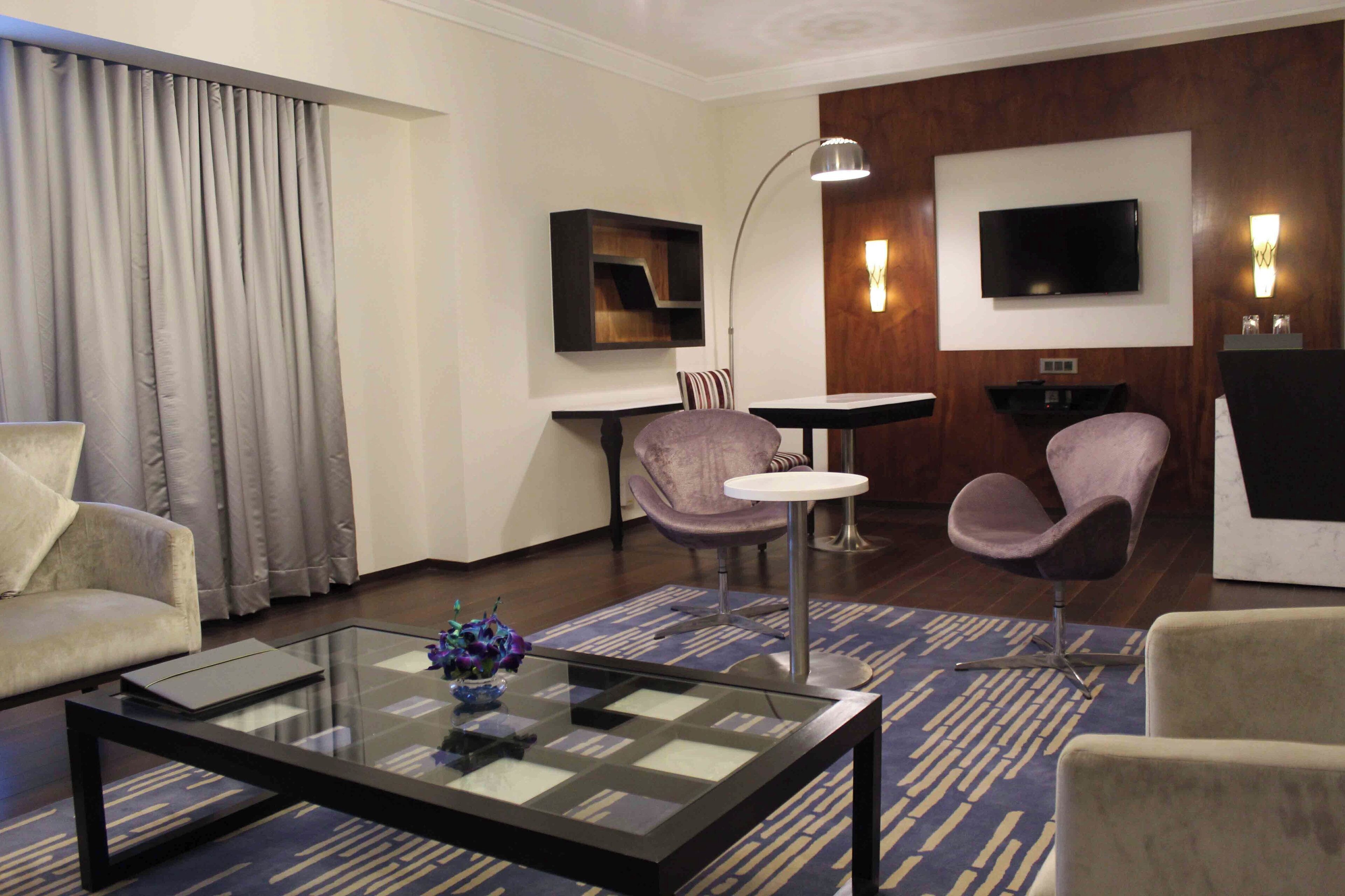 Une chambre d’hôtel moderne avec une table basse en verre, deux fauteuils, un canapé, un téléviseur à écran plat et une petite table avec une lampe.