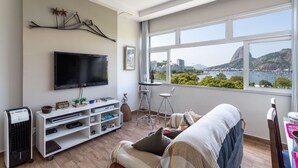 A modern living room with a flat-screen TV, a white entertainment unit, a small table, two chairs, a sofa, and a large window offering a city view. - Moderno Apartamento na Praia de Botafogo (Rio de Janeiro)