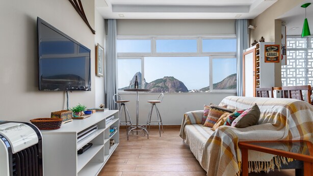A modern living room with a large TV, a comfortable sofa, a small dining area, and a view of a mountain. - Moderno Apartamento na Praia de Botafogo (Rio de Janeiro)