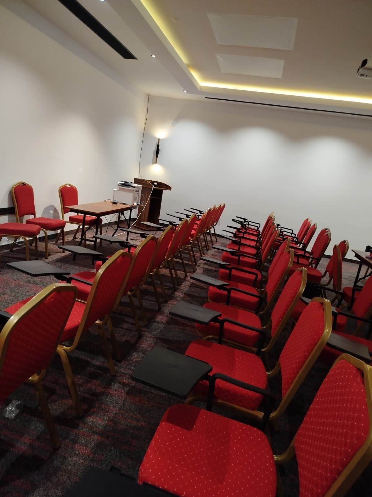 Un salón de conferencias con filas de sillas rojas y mesas, un atril y suelo alfombrado.