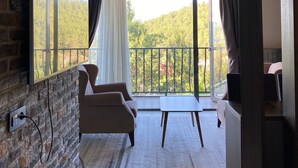 A modern room with a stone wall, a flat-screen TV mounted on it, a sofa, a small table, and a balcony with a view of trees. - Amorium Hotel (Kastamonu)