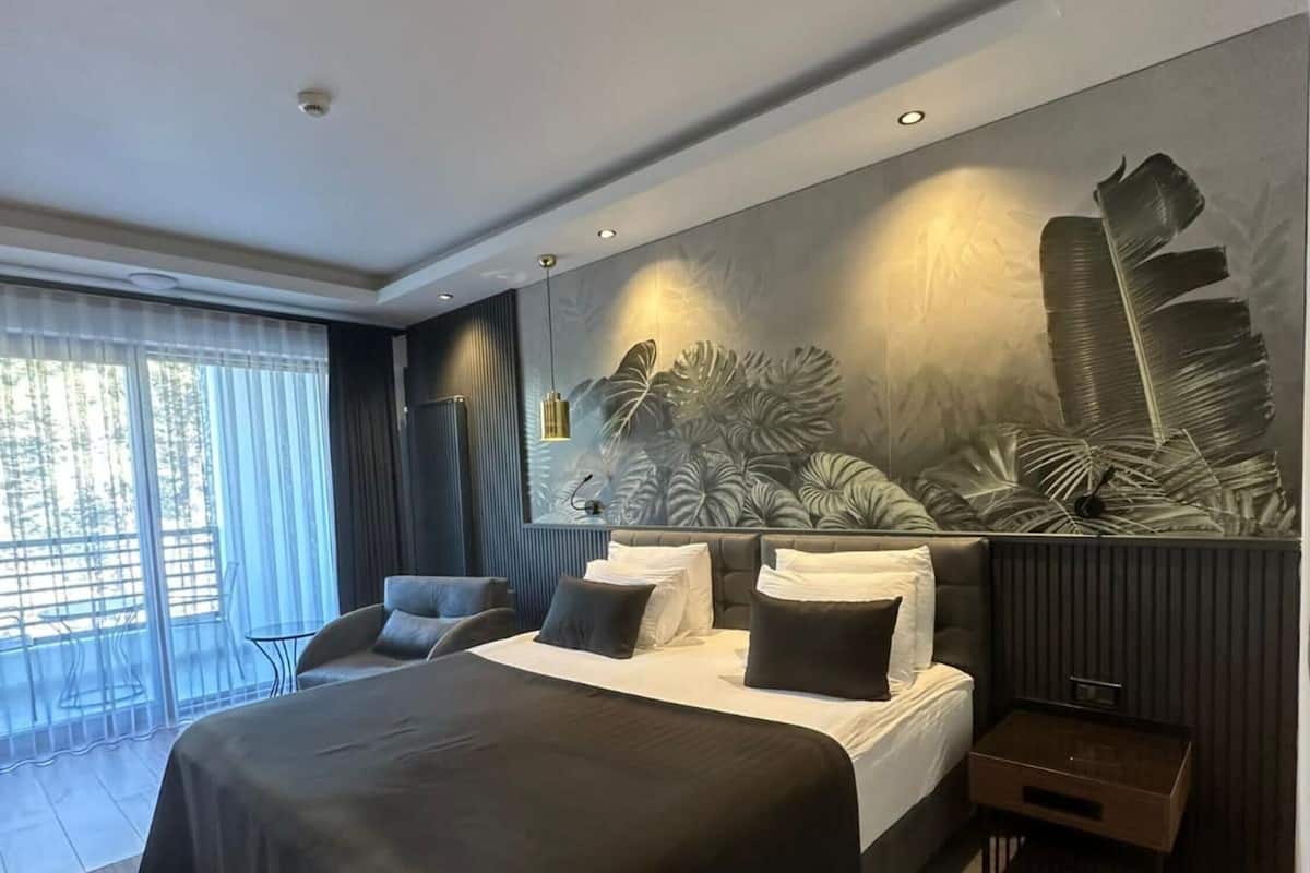 A modern bedroom with a large bed, a seating area with a chair, a small table, and a view of the outdoors through large windows.