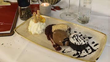 Un plato de postre con mousse de chocolate, crema batida y un pastel espolvoreado de chocolate, acompañado de una vela y un vaso sobre una mesa.
