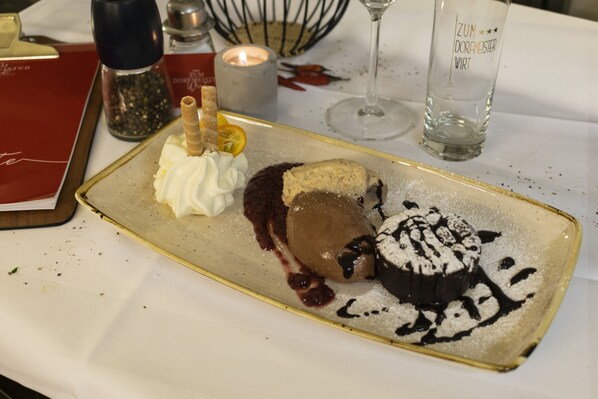 A dessert plate with chocolate mousse, whipped cream, and a chocolate-dusted cake, accompanied by a candle and a glass on a table.