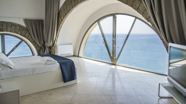 A bedroom with a bed, a TV, and a view of the sea through large arched windows. - Sunrise Guest House (Gaeta)