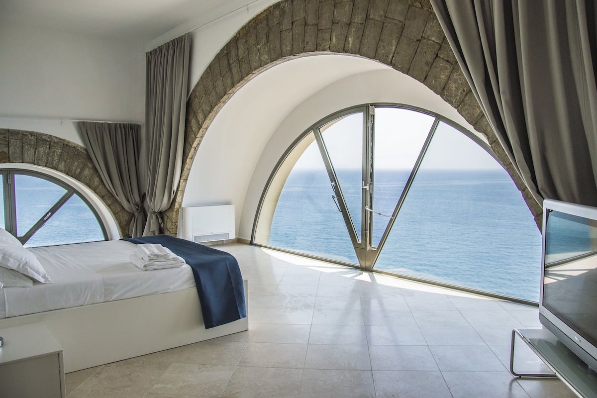 Quarto com cama, TV e vista para o mar através de amplas janelas em arco.