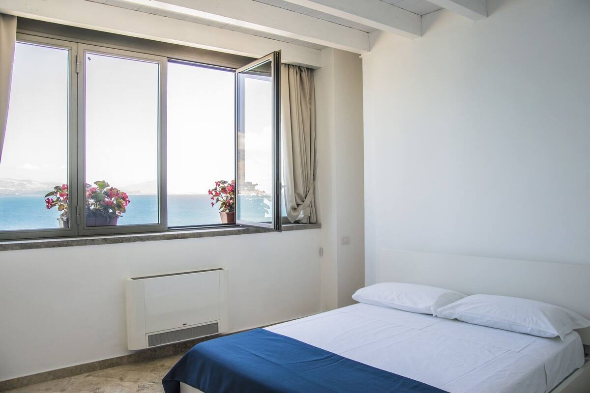 Quarto com cama, janela com cortinas e vista para o mar.
