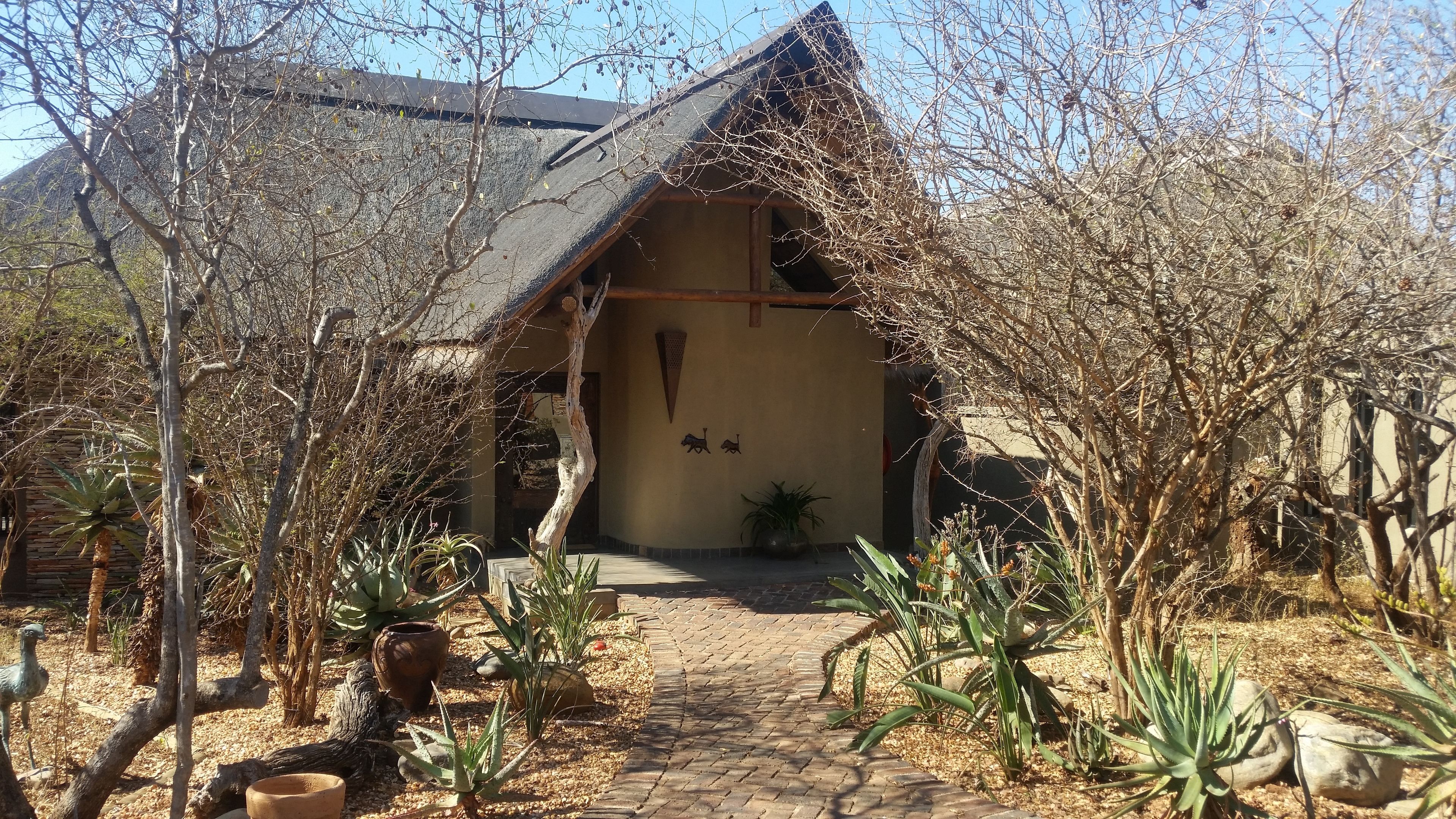 A rustic building with a thatched roof, surrounded by dry vegetation and a paved pathway leading to the entrance.