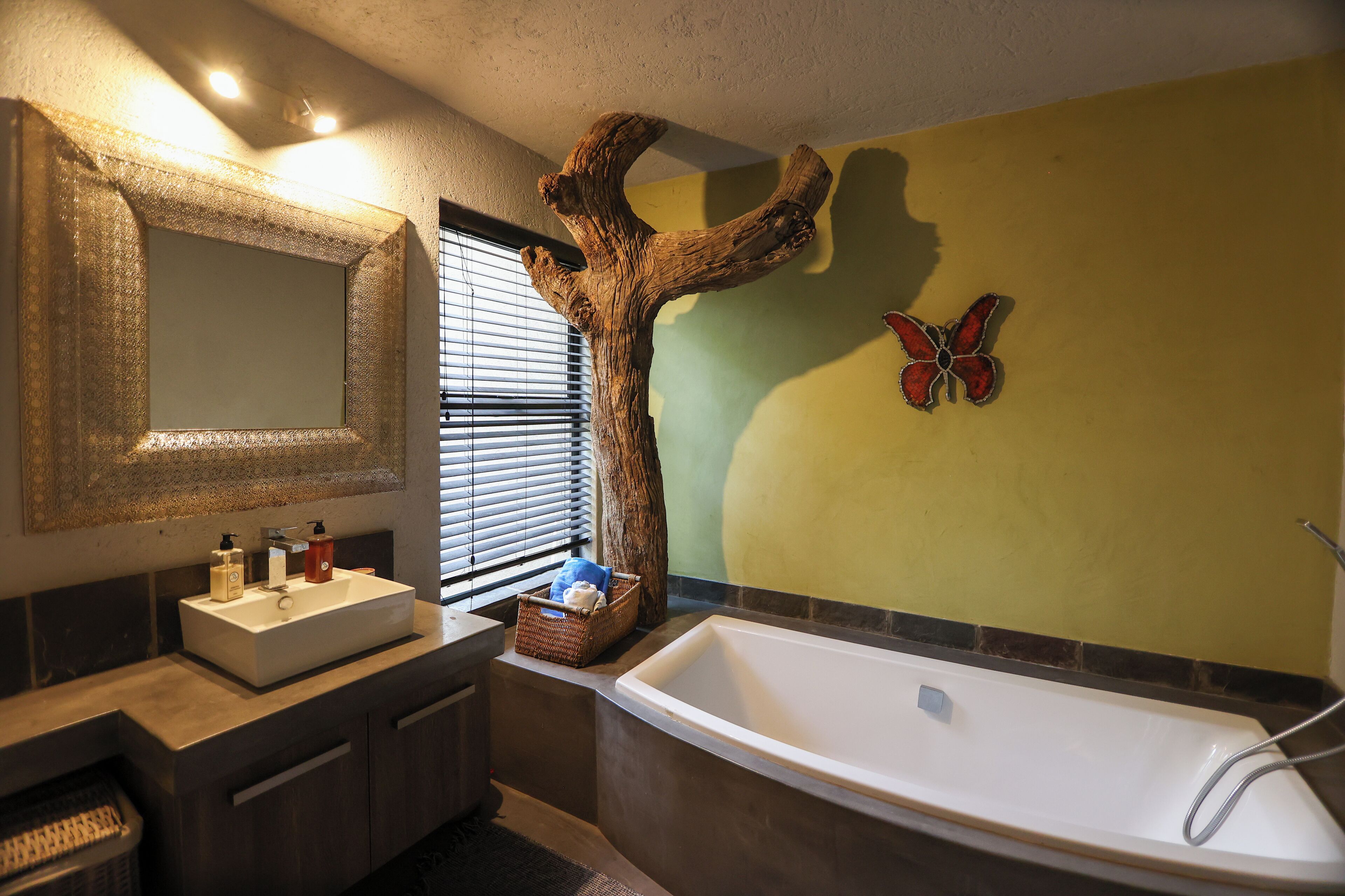 A bathroom with a large mirror, a wooden tree sculpture, a butterfly decoration, a bathtub, and a window with blinds.