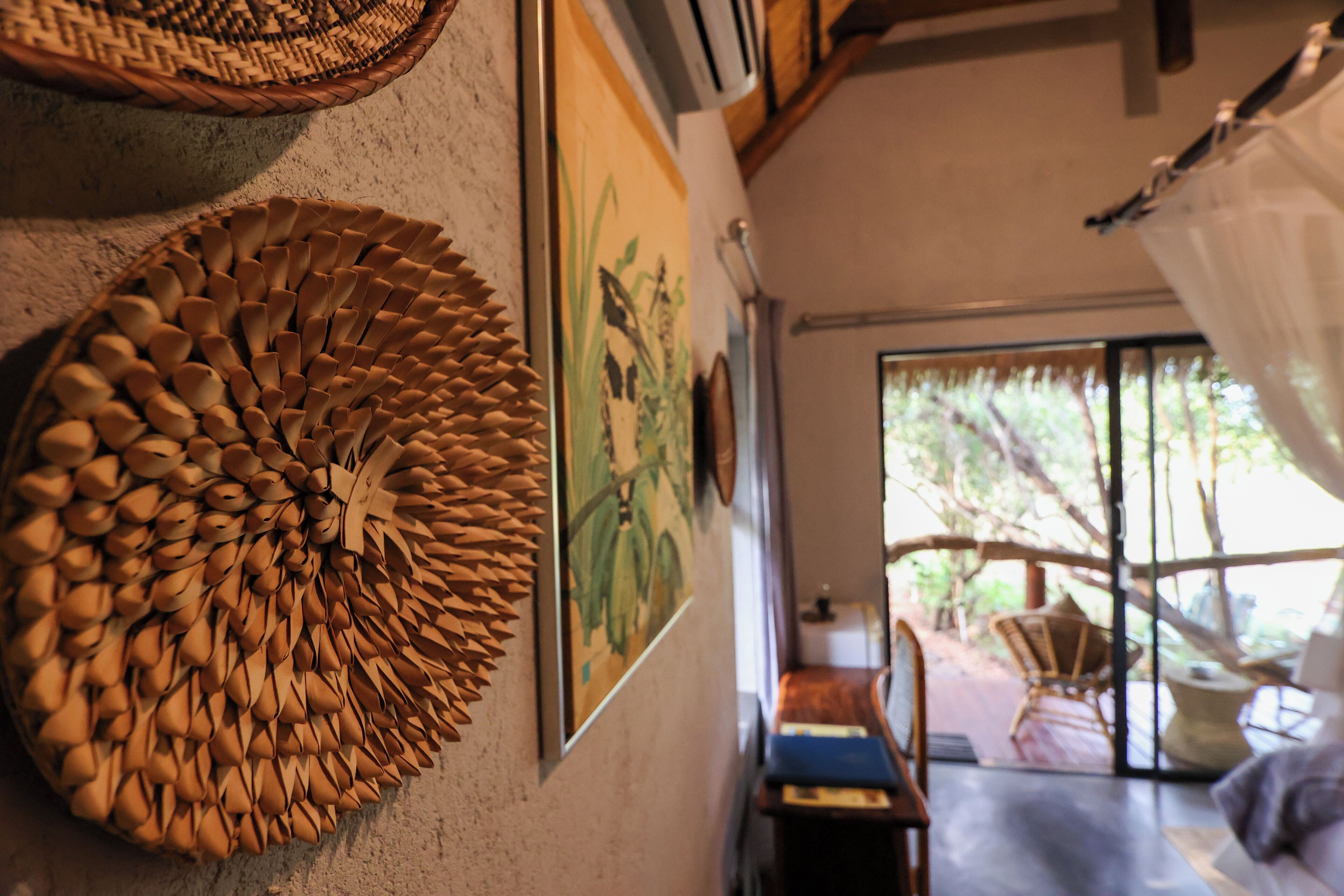 A room with a woven wall decoration, a framed painting, and a view of an outdoor area with furniture.