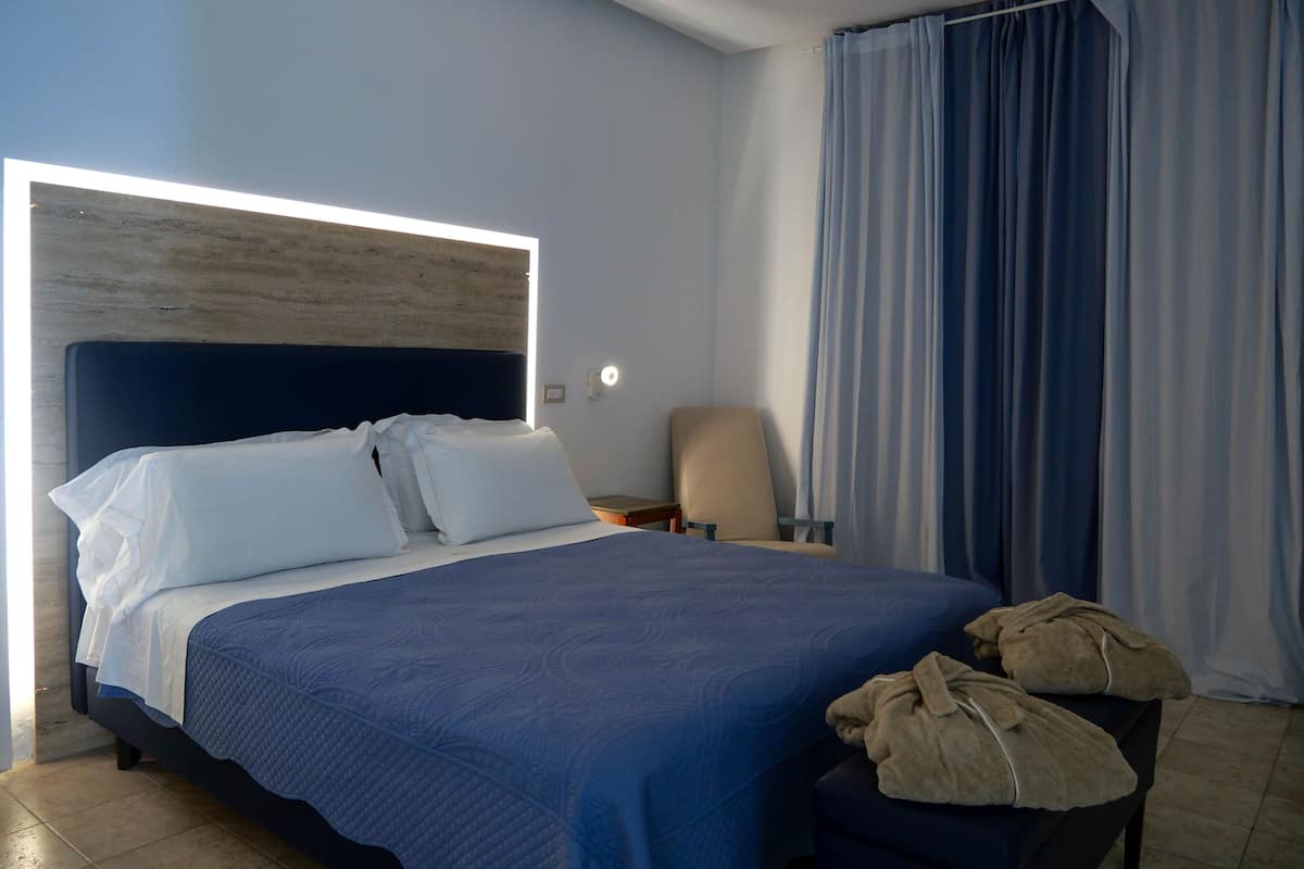 A modern hotel room with a large bed, a headboard with built-in lighting, a chair, and a window with curtains.