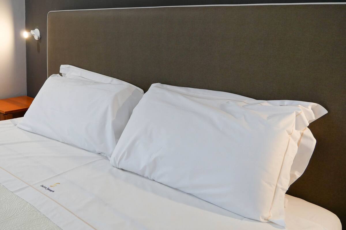 A neatly made bed with white bedding and pillows, a wooden nightstand, and a wall-mounted light fixture.