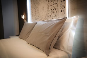 En säng med beige sängkläder och kuddar, ett träfärgat sänggavel och en mönstrad vägg bakom.