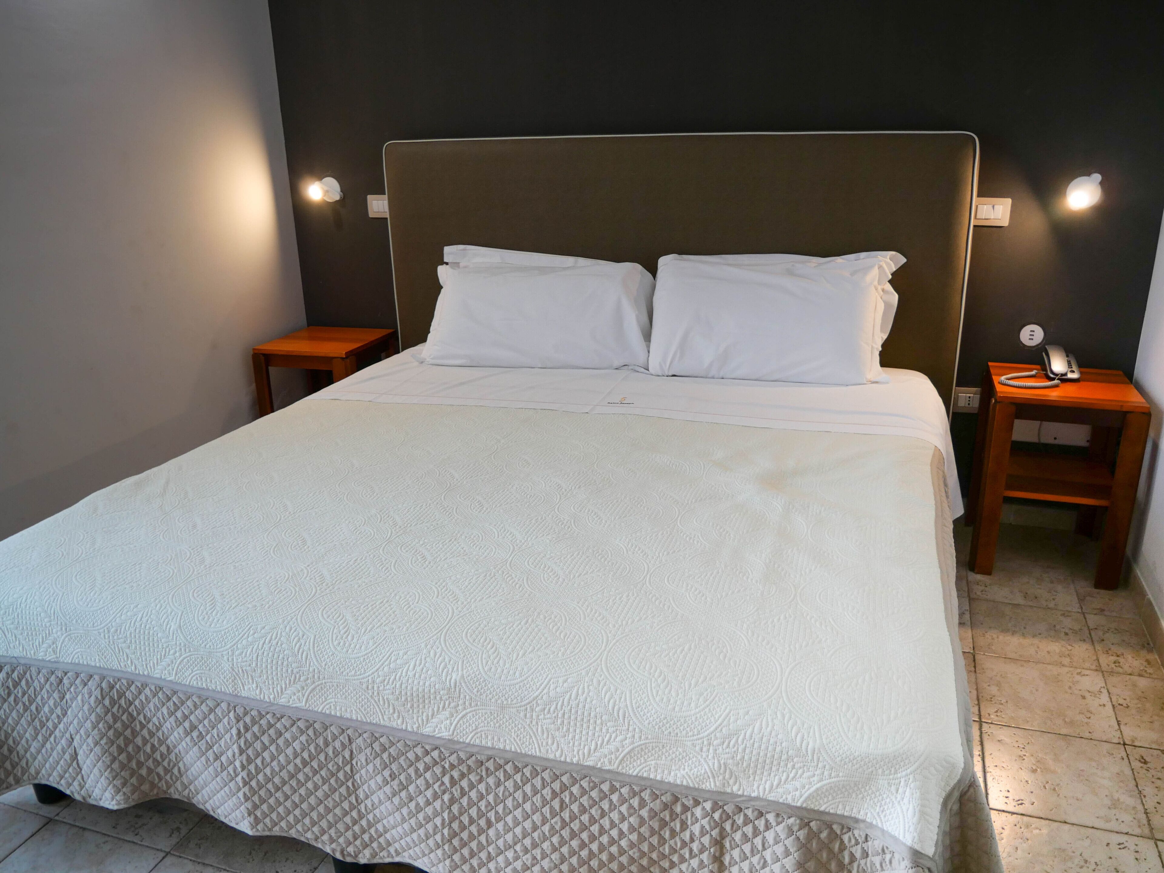A neatly made bed with a quilted bedspread and two pillows, flanked by wooden nightstands with a telephone on one.