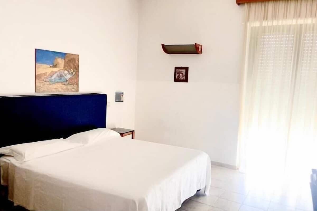 A hotel room with a large bed, a nightstand, a painting on the wall, and a window with curtains.