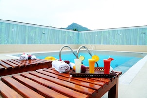 Zona junto a la piscina con tumbonas de madera, una bandeja con toallas, frutas y bebidas.