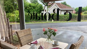 Une table dressée pour deux personnes, avec de la vaisselle à motifs floraux, un vase rempli de fleurs roses, et une vue sur une maison, visible à travers une fenêtre sur laquelle on peut lire le mot « BORG ».