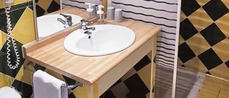 Un baño con lavamanos blanco, espejo e inodoro. Hay una zona de ducha con puerta de vidrio y una toalla colgada en la pared.