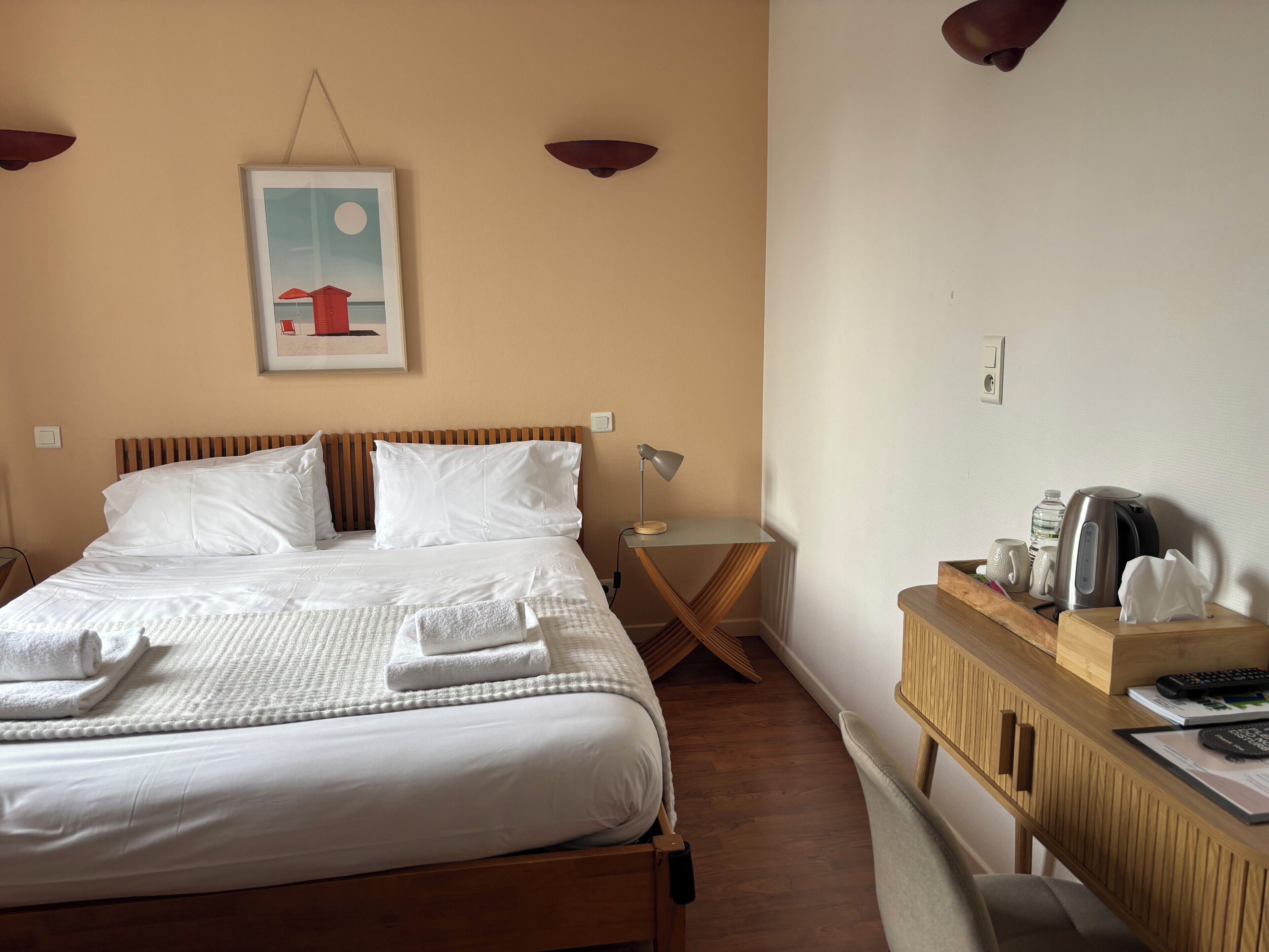 A neatly made bed with white linens, a bedside table with a lamp, a small wooden desk with a kettle, and a framed picture on the wall.