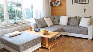 A cozy living room with a sectional sofa, a small table with a fruit bowl, and a bookshelf. - Hus Mattgoot (St. Peter-Ording)