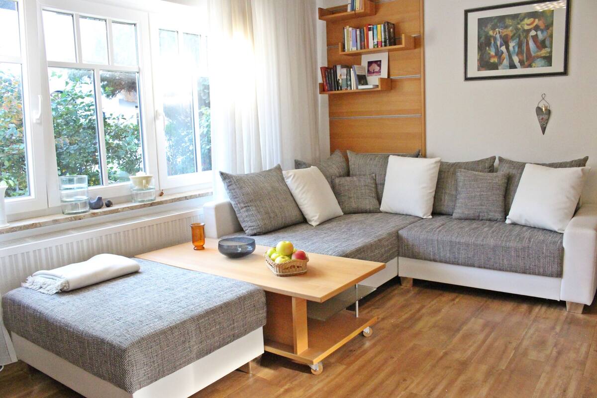 A cozy living room with a sectional sofa, a small table with a fruit bowl, and a bookshelf.